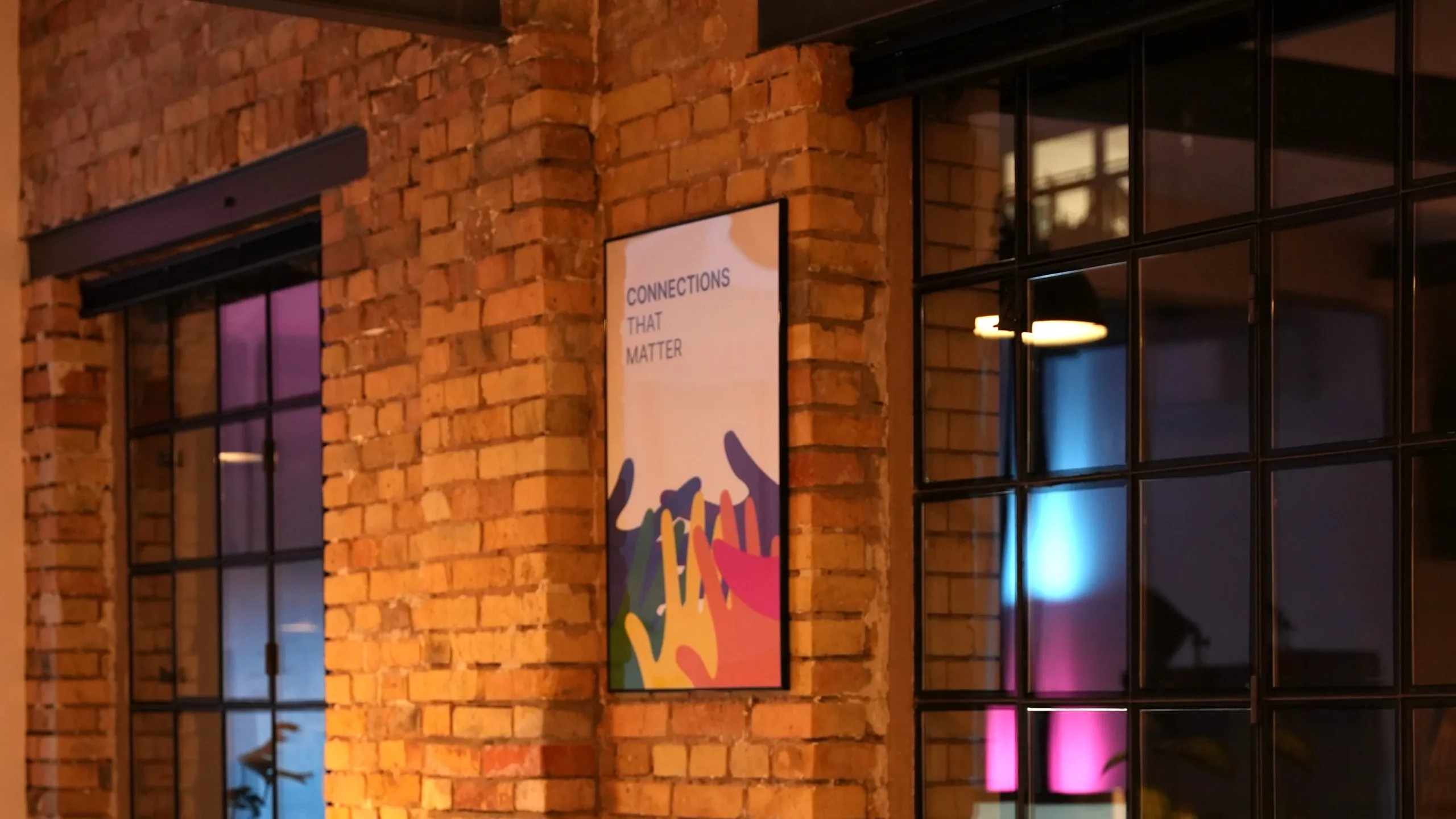 Ein vertikales Poster mit der Aufschrift "CONNECTIONS THAT MATTER" und einer bunten Illustration von ineinandergreifenden Händen hängt an einer rustikalen Backsteinwand. Die Szene befindet sich in einem Innenraum mit großen, dunkel gerahmten Sprossenfenstern, durch die im Hintergrund stimmungsvolle lila und blaue Lichteffekte einer Lichtshow zu sehen sind.