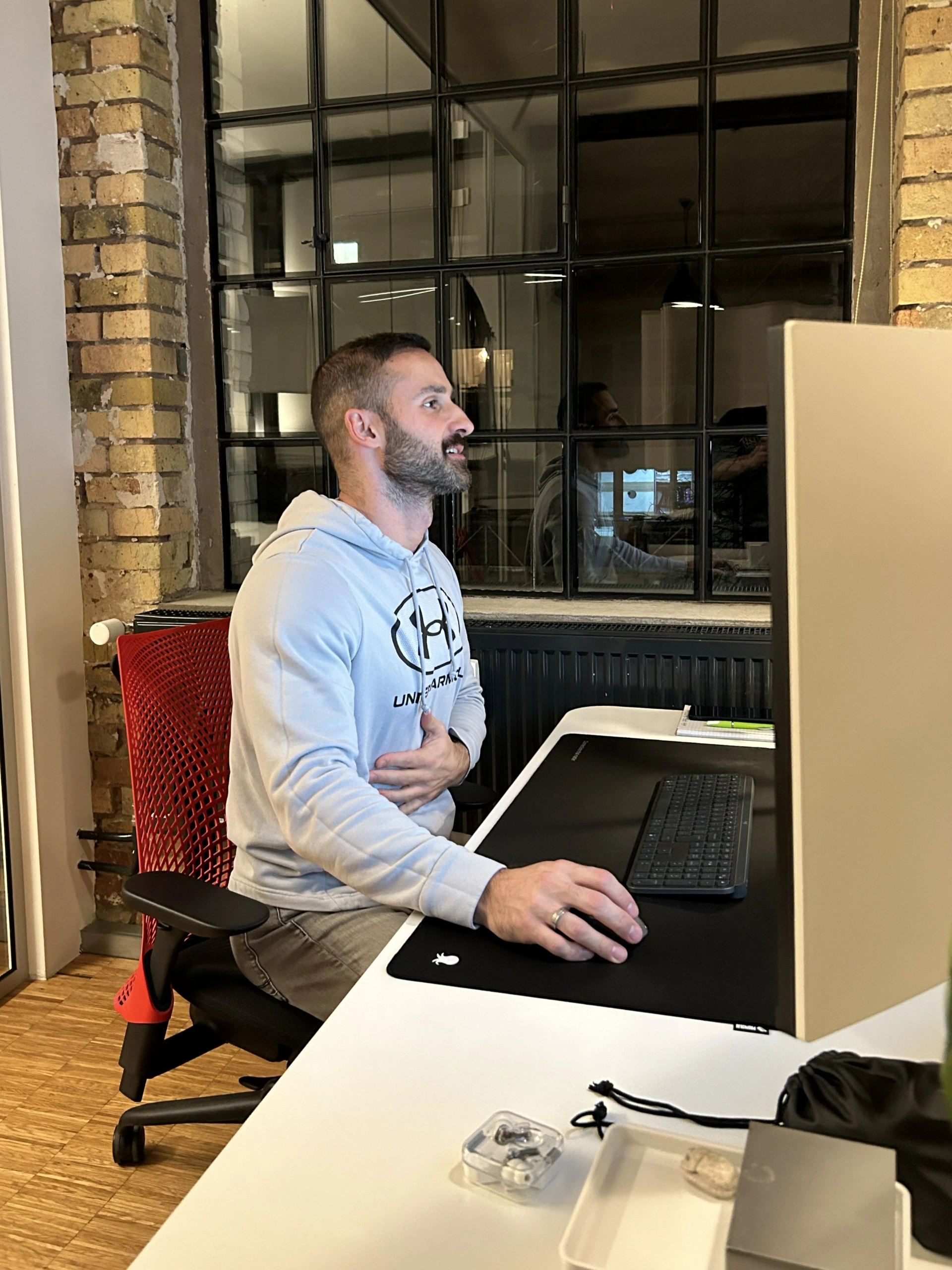 Ein Mann mit Bart sitzt im Profil an einem weißen Schreibtisch und arbeitet an einem Computer. Er trägt einen hellgrauen Kapuzenpullover der Marke Under Armour und blickt lächelnd auf den Monitor, der nur von der Rückseite zu sehen ist. Er sitzt auf einem Bürostuhl mit einer markanten roten Netz-Rückenlehne. Im Hintergrund befindet sich eine rustikale Backsteinwand mit einem großen, dunkel gerahmten Sprossenfenster, in dem sich der Innenraum spiegelt.