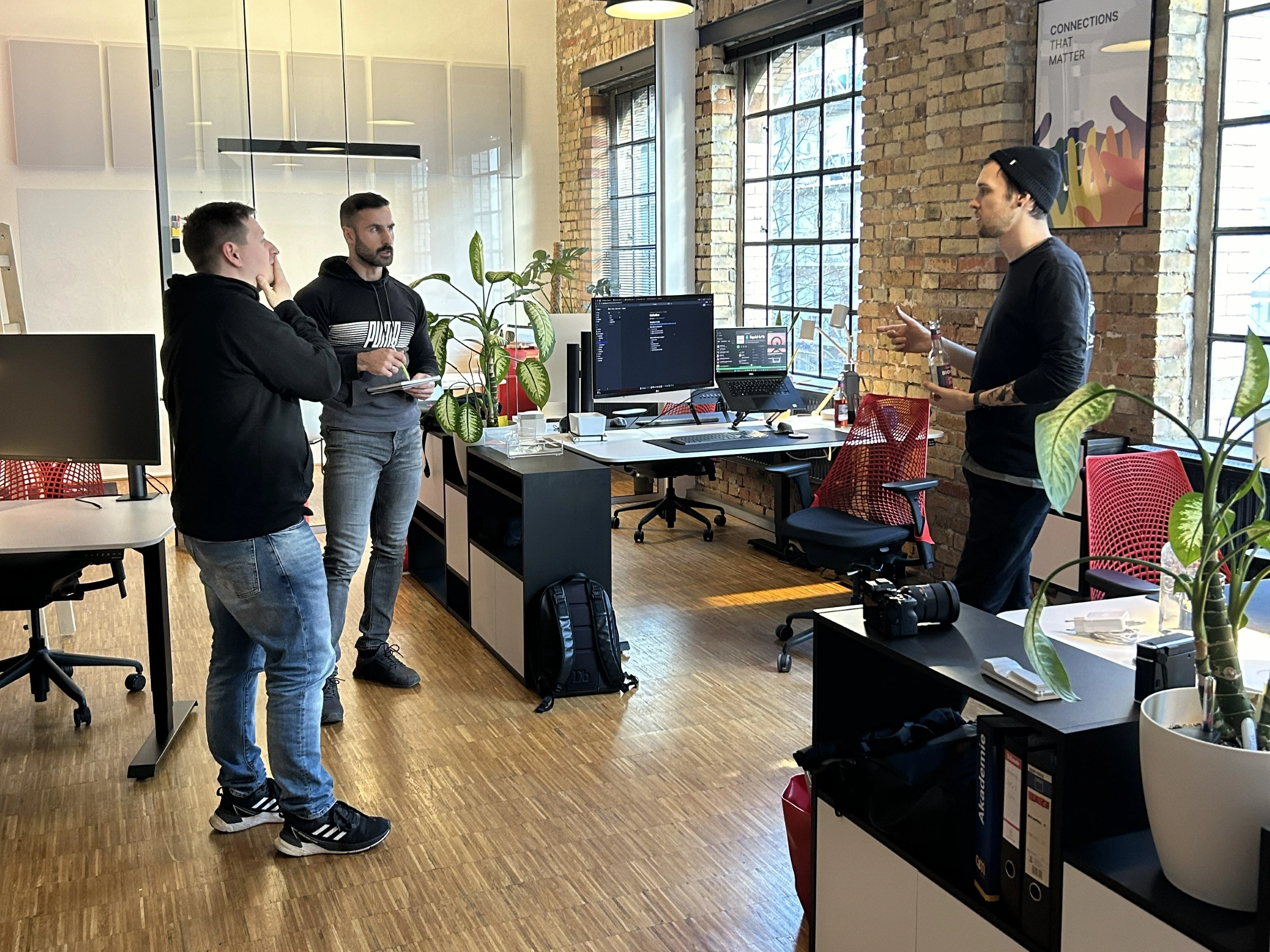 Drei Männer stehen in einem hellen Loft-Büro mit Backsteinwänden und großen Sprossenfenstern und führen ein Gespräch. Ein Mann im Vordergrund links trägt einen schwarzen Hoodie und Jeans, während ein anderer Mann in der Mitte ein Notizbuch hält. Rechts steht ein dritter Mann mit schwarzer Beanie und hält eine Getränkeflasche. Im Hintergrund sind moderne Arbeitsplätze mit Monitoren, ergonomischen Stühlen mit auffälligen roten Rückenlehnen sowie mehrere große Zimmerpflanzen zu sehen.