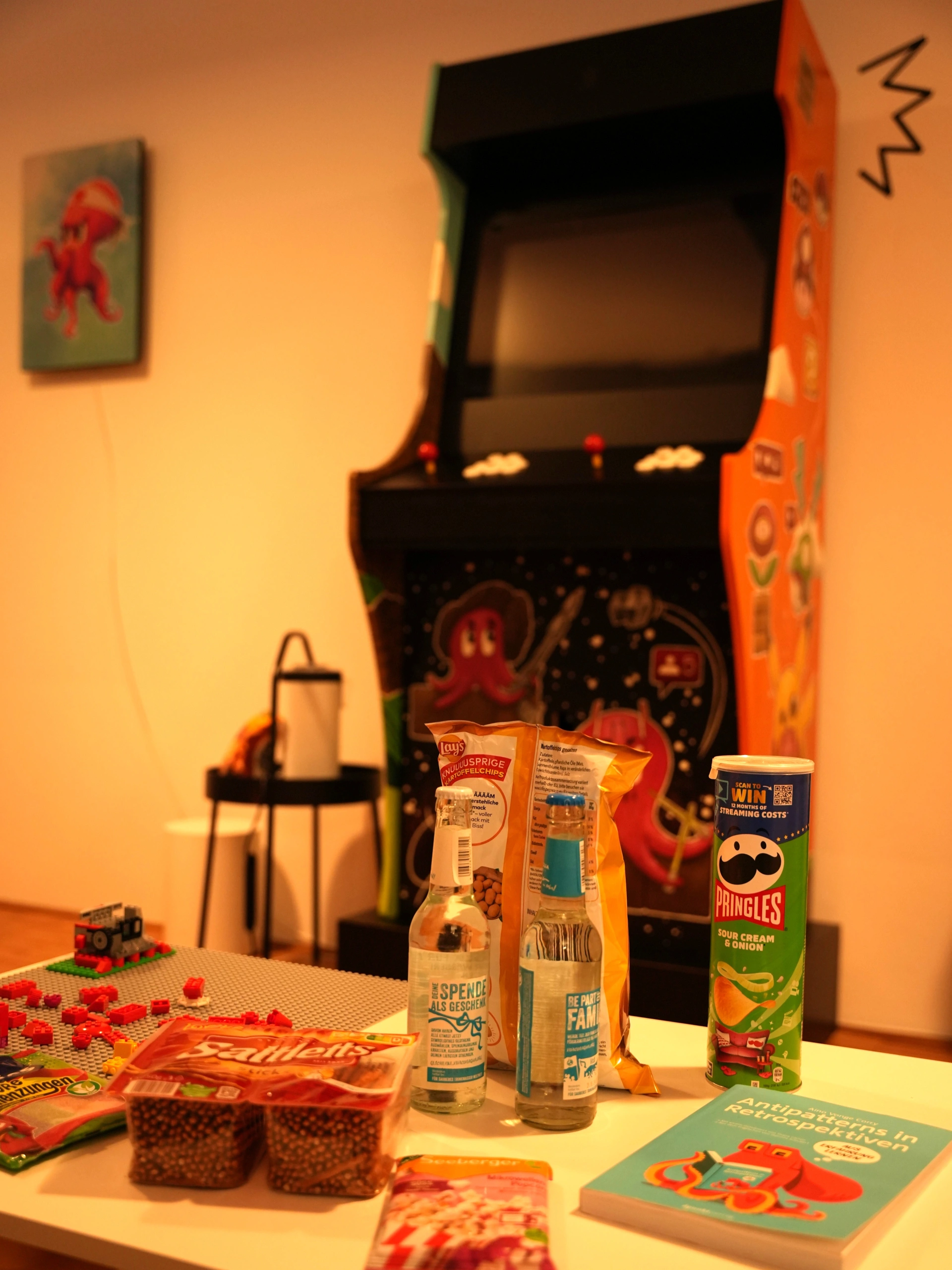 Ein heller Wohnraum mit einem weißen Tisch im Vordergrund, auf dem verschiedene Snacks und Getränke verteilt sind: eine Packung Salzstangen, eine Dose Pringles, zwei Glasflaschen, eine Chipstüte und ein Buch mit dem Titel „Antipatterns in Retrospektiven“. Im unscharfen Hintergrund steht ein großer, bunt verzierter Arcade-Spielautomat neben einer Wand, an der ein Bild mit einem roten Oktopus-Motiv hängt. Links auf dem Tisch sind zudem rote LEGO-Steine auf einer grauen Bauplatte zu sehen.
